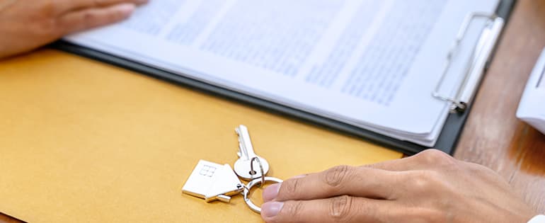 Mão masculina segurando ma chave com chaveiro de uma casa prateado em uma mesa com contrato de reajuste de aluguel